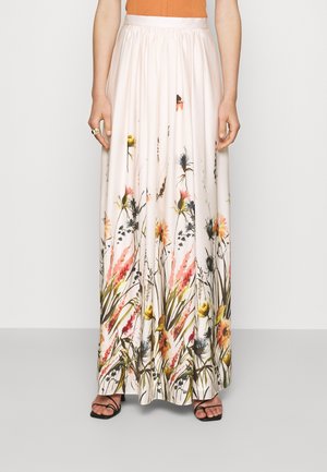 Femme portant une longue jupe blanche avec un imprimé floral coloré à l'ourlet, associée à des sandales noires à lanières.