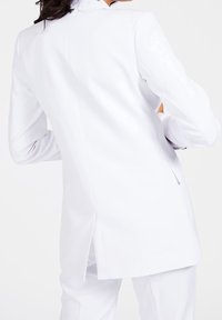 Valkoinen räätälöity takki, jossa on korkea kaulus, yksi takatuuletin, jämäkät olkapäät ja sileä pinta. Täydentää virallista pukeutumista.