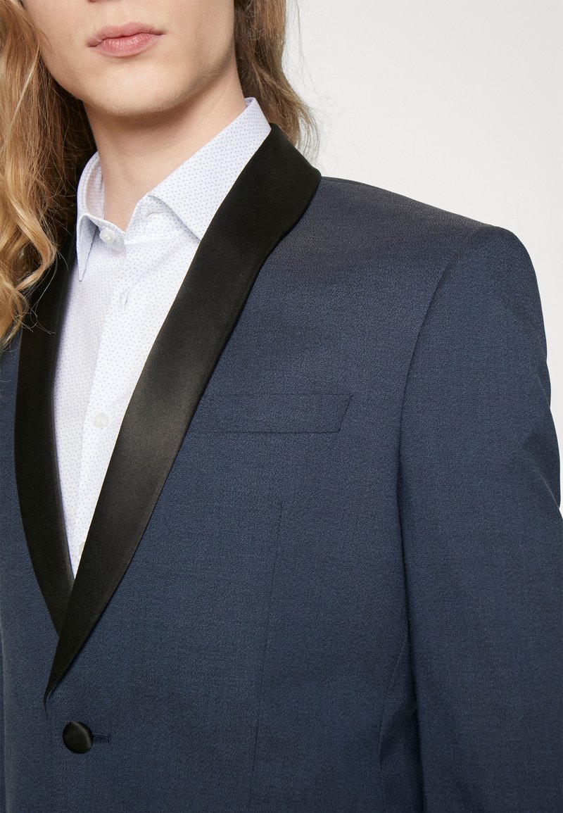 Giacca smoking blu navale con colletto a scialle in satin nero e una piccola tasca sul petto. Sotto, una camicia da dress leggera con un motivo è parzialmente visibile.