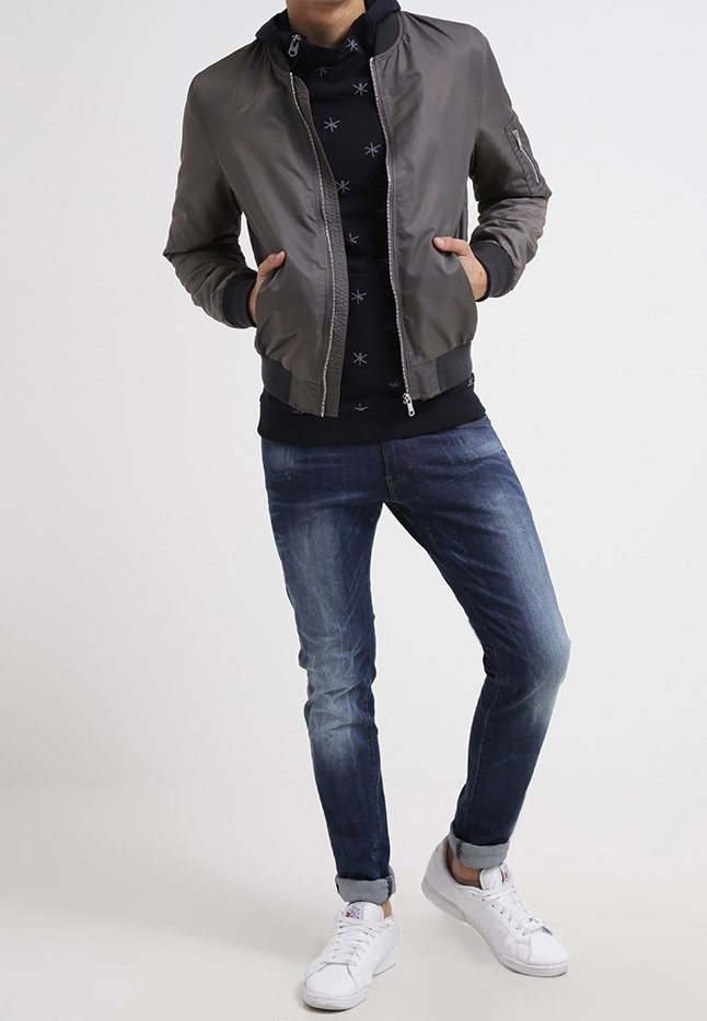 Šedá bomber bunda přes černou mikinu se vzorem hvězd, spárovaná s modrými džíny a bílými teniskami. Hladká tkanina, neformální design.