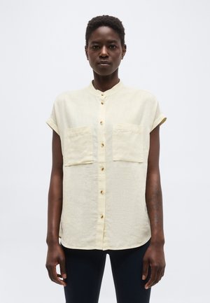 Personne portant une chemise beige à manches courtes, boutonnée, avec deux poches sur la poitrine, debout devant un fond blanc uni.