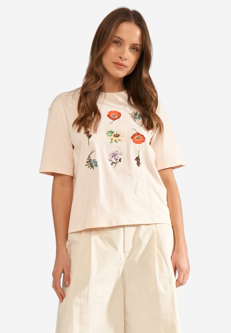 Femme portant une chemise beige à manches courtes avec des fleurs brodées colorées et un pantalon beige ample assorti, debout devant un fond blanc.