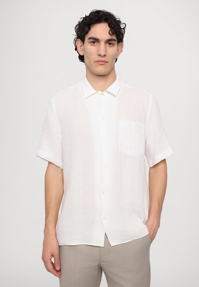 Camisa de lino blanca de manga corta con cuello abotonado, bolsillo frontal y corte relajado, combinada con pantalones gris claro.