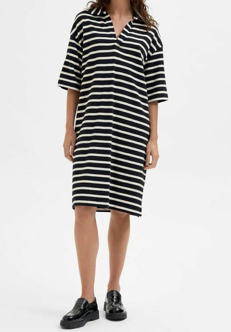 Femme portant une robe noire et blanche à rayures horizontales, aux genoux, à manches courtes, et des mocassins noirs, se tenant devant un fond blanc.