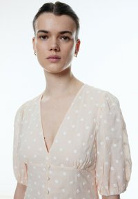 Hellbeige Kleid mit weißen Punkten, tiefem V-Ausschnitt, kurzen Puffärmeln und Knopfdetails vorne. Texturiertes Material, lässiges Design.