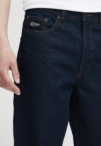 Jeans de mezclilla azul oscuro con costuras naranjas, que presentan un bolsillo frontal con una etiqueta. La tela tiene una textura suave y un diseño clásico.