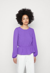 Blusa longa de mangas com decote redondo, em tom de roxo, com tecido texturizado, cintura franzida e mangas bufantes, combinada com calças brancas.