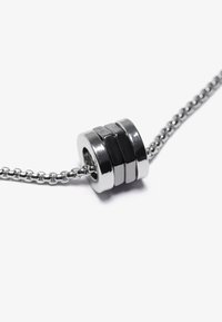 Metallkjede med en sylinderformet medaljong som har vekslende svarte og sølvfarvede seksjoner, samt en perlekjede. Polert, moderne design.