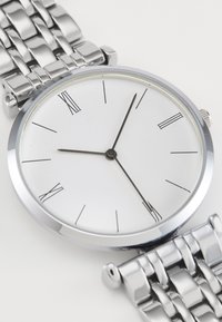 Silberne Metall-Uhr mit rundem Zifferblatt, weißem Ziffernblatt, römischen Ziffern und schwarzen Zeigern. Verfügt über ein massives Gliederarmband. Minimalistisches Design.