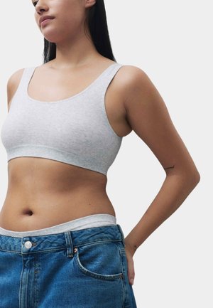 Giovane donna che indossa un top grigio corto e jeans azzurri, con le mani appoggiate sui fianchi su uno sfondo semplice.