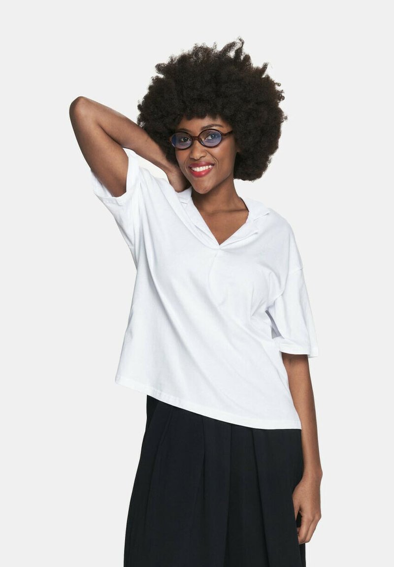 Biała, oversize'owa koszulka z dekoltem w kształcie litery V i krótkimi rękawami, noszona z czarną plisowaną spódnicą, na jednolitym tle.