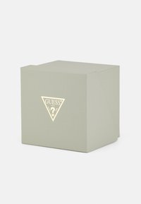 Kvadratinė dovanų dėžutė šviesiai žaliame tekstūruotame popieriuje, su auksiniu trikampiu logotipu, kuriame centre yra žodis "GUESS". Paprastas, elegantiškas dizainas.