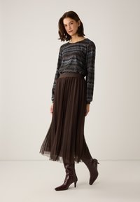 Langer brauner Tüllrock kombiniert mit einem schwarz-blau gemusterten Langarmshirt. Das Modell trägt glänzende burgunderrote Stiefel mit Absatz.