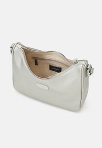 Petit sac à bandoulière argenté avec fermeture éclair, intérieur beige, poche zippée intérieure et étiquette de marque en métal à l'avant.