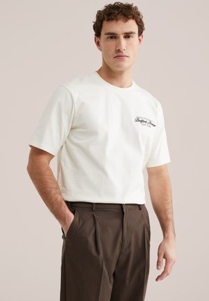 Göndör hajú fiatal férfi fehér rövid ujjú pólót visel "Bedford Avenue New York" felirattal, barna nadrágban, egyik kezét a zsebében tartva áll.