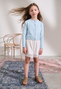 Light blue knitted cardigan with button closure, paired with a white skirt featuring lace trim. Brown strappy sandals; wavy hair.