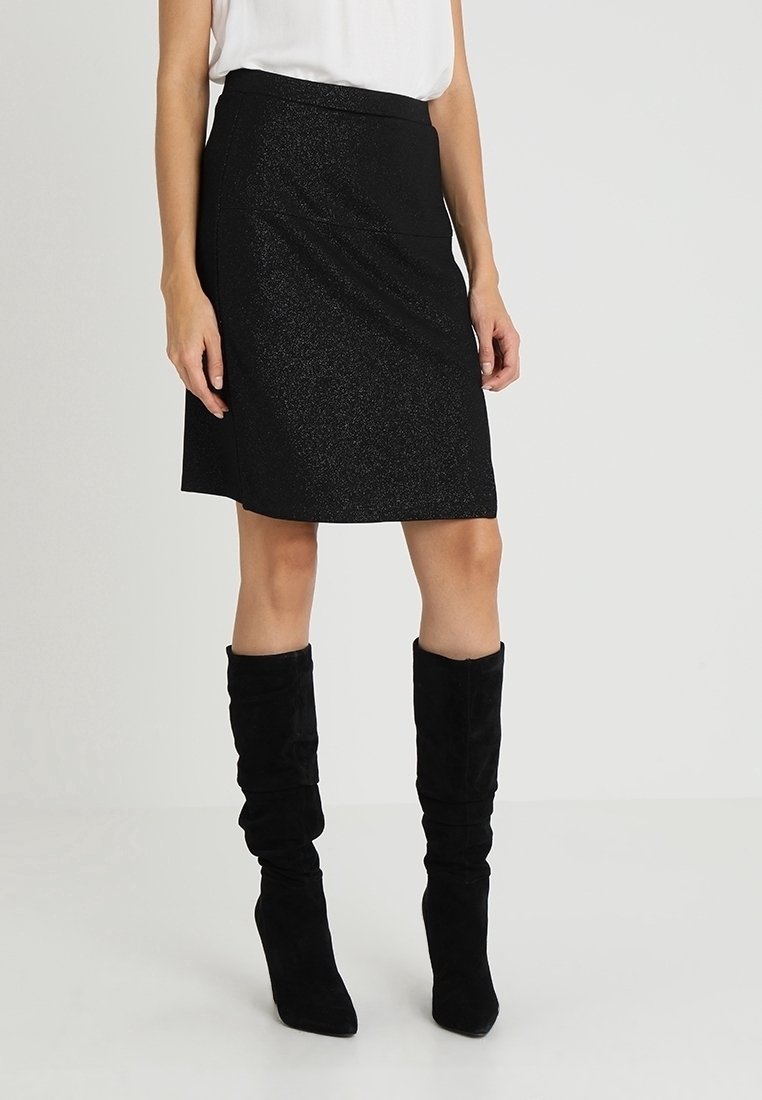 Femme portant une jupe noire mi-genou et des bottes hautes noires décontractées, debout devant un fond clair uni.