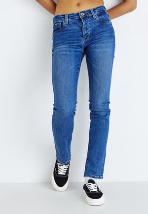 Personne portant un jean skinny bleu taille haute et des baskets basses noires avec des lacets blancs, debout devant un fond blanc.