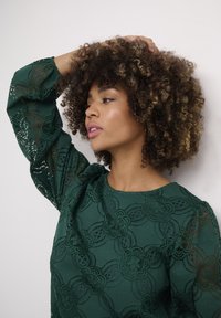 Grön spetsblus med blommönster och halvgenomskinliga ärmar. Modellen poserar med lockigt hår, handen på huvudet, mot en neutral bakgrund.