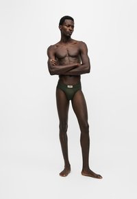 Homme torse nu à la peau foncée portant un slip vert foncé, debout pieds nus, bras croisés, regardant légèrement sur le côté sur un fond blanc uni.