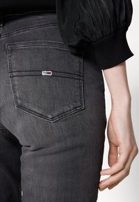 Gros plan sur une personne portant un jean en denim noir avec une poche arrière ornée d'un petit logo rectangulaire rouge, blanc et bleu, et un top noir à manches bouffantes.