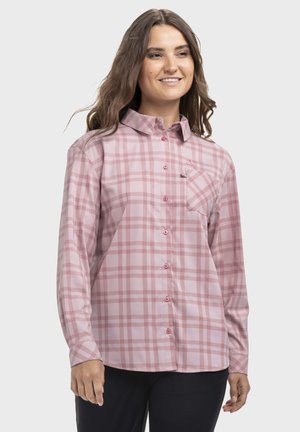 Frau lächelt, trägt ein pink kariertes, langärmliges Hemd mit Knöpfen, Kragen und Brusttasche, steht vor einem weißen Hintergrund.