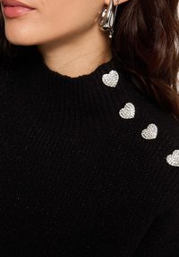 Suéter negro de punto con un cuello alto, que presenta cinco apliques de corazones de strass brillantes a lo largo del hombro. Textura suave, silueta ajustada.