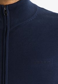 Mörkblå zip-up tröja med ribbat krage, tillverkad av ett slätt stickat material. Har subtila logotyper nära fållen.