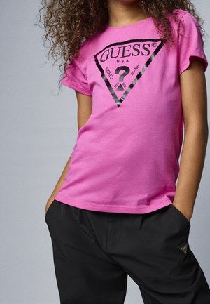 Enfant portant un t-shirt rose Guess avec logo et pantalon noir, mains dans les poches, cheveux bouclés visibles, debout devant un fond uni.