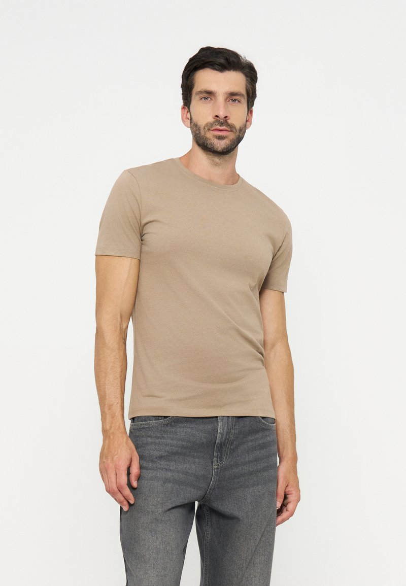 Man met donker haar en baard, gekleed in een beige T-shirt met korte mouwen en grijze spijkerbroek, staand tegen een effen witte achtergrond.