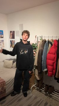 Νέος άνδρας που φορά μαύρο φούτερ με κουκούλα στέκεται στο υπνοδωμάτιο δίπλα στο κρεβάτι, σε κρεμάστρα με μπουφάν και σε έναν τοίχο με φωτογραφίες και φυτά.