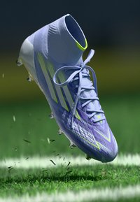 Svetlofialová futbalová kopačka s vysokým golierom, so žltými akcentmi, štruktúrovaným zvrškom a kopačkovou podrážkou, suspendovaná nad trávou.