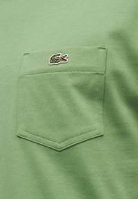 Πράσινη υφασμάτινη τσέπη στο στήθος σε πουκάμισο με ένα μικρό κεντητό πράσινο και λευκό λογότυπο κροκόδειλου πάνω της.