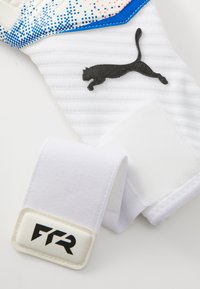 Vita målvaktsvante med texturerad greppyta, blå accenter och svart Puma-logotyp. Säkerhetsremmen har vit kardborrestängning och varumärkeslogotyper.
