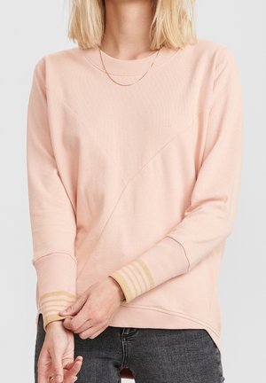 Žena nosící světle růžový dlouhý rukáv s béžovými proužkovanými manžetami a tmavými džíny, upravuje jeden rukáv s dlaněmi sepnutýma před sebou.