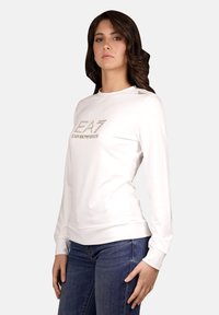 Weißer Pullover aus glattem Stoff, mit einem metallischen Logo-Print auf der Vorderseite, langen Ärmeln und gerippten Bündchen. Jeans sind darunter sichtbar.