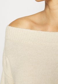 Mjuk beige off-shoulder tröja med ribbad halsringning och texturerat stickat mönster. Tyget verkar lätt och ventilerat.