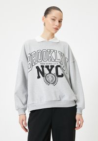 Sudadera gris con cuello blanco, que presenta el texto impreso en negro "BROOKLYN NYC" y un diseño de emblema. Corte relajado con puños acanalados.