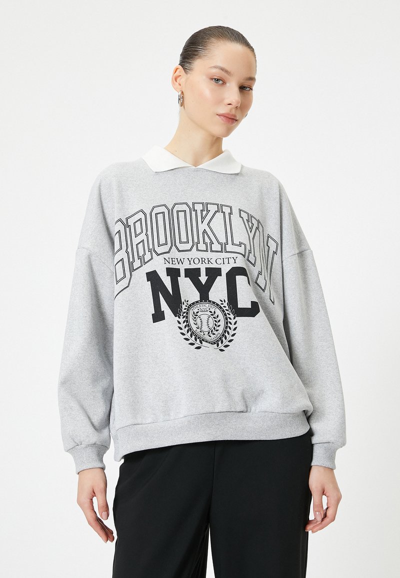Sudadera gris con cuello blanco, que presenta el texto impreso en negro "BROOKLYN NYC" y un diseño de emblema. Corte relajado con puños acanalados.