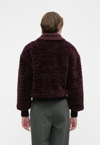 Osoba viditeľná zozadu, oblečená v tmavo purpurovej štruktúrovanej fleecovej bunde s rebrovaným golierom a manžetami, v kombinácii so sivými nohavicami na jednofarebnom pozadí.