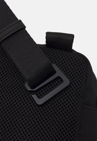 Zaino in rete nera, angolo che mostra le cinghie in nylon resistenti e un attacco con fibbia in plastica. Superficie testurizzata con un bordo rinforzato.