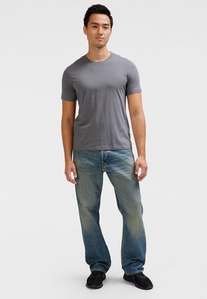 Grå t-shirt med korte ærmer, afslappet pasform, parret med slidte blå jeans. Fodtøj: sorte sneakers. Ensfarvet baggrund, ingen mønstre eller logoer.