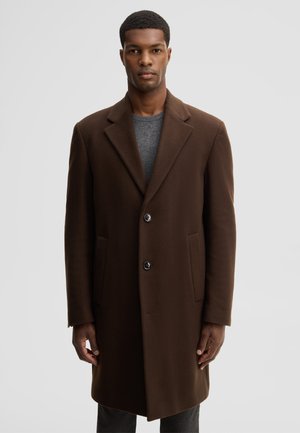 Man wearing a dark brown buttoned coat over a gray sweater, standing against a plain light gray background.