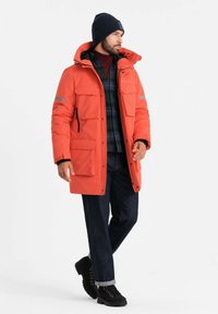 Heldere oranje pufferjack met reflecterende accenten, meerdere voorzakken en een capuchon, gecombineerd met donkere jeans en zwarte veterschoenen.