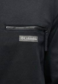 Μαύρο ύφασμα με οριζόντια τσέπη με φερμουάρ και ραμμένη γκρι ετικέτα της μάρκας Columbia κάτω από το φερμουάρ.