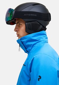 Zwarte skihelm met een reflecterende groene lens, gedragen met een blauwe jas met een hoge kraag en zichtbare rits. Logo op het helmriempje.