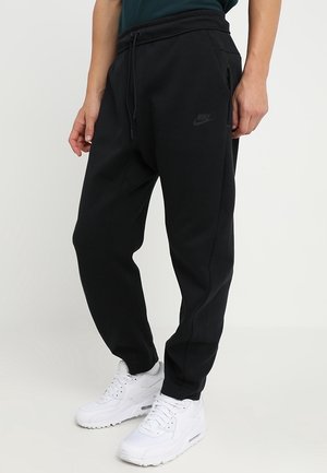 Person trägt schwarze Nike-Jogginghosen mit Kordelzugbund und Reißverschlusstasche, kombiniert mit weißen Sportschuhen, steht auf einer ebenen Fläche.