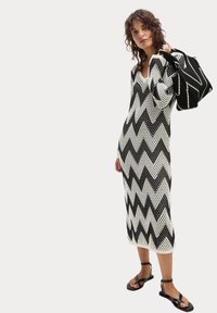 Un vestito a maglia chevron bianco e nero presenta una silhouette aderente, scollo a V e maniche lunghe, abbinato a una borsa con motivo a zigzag coordinato e sandali.