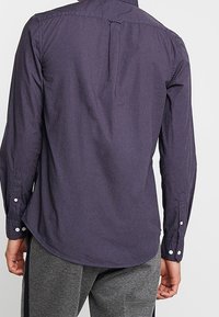 Camicia a maniche lunghe blu navy con colletto a punta, plissé posteriore e orlo arrotondato. Presenta polsi con bottoni e una texture di tessuto liscia.