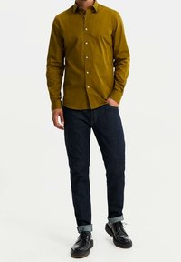 Sinappikeltainen napitettava paita, jossa on terävä kaulus, yhdistetty tummansinisistä farkuista käännettyihin lahkeisiin ja mustat nauhalliset kengät.